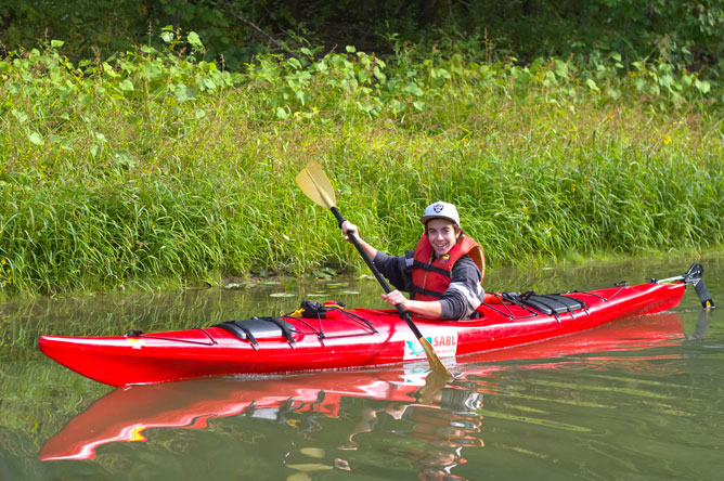 Side view of a young man paddling a red kayak along a channel.