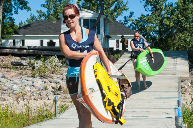 Two women carrying their sailboards on a dock.